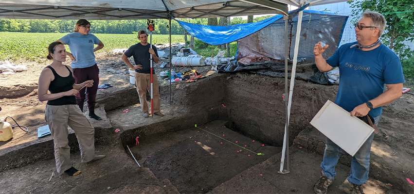 Archeologists standing in an excavated hole under a canopy